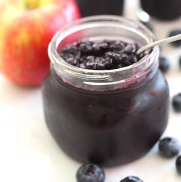 A jar of Blueberry Apple Jam, shown next to fresh blueberries and an apple.