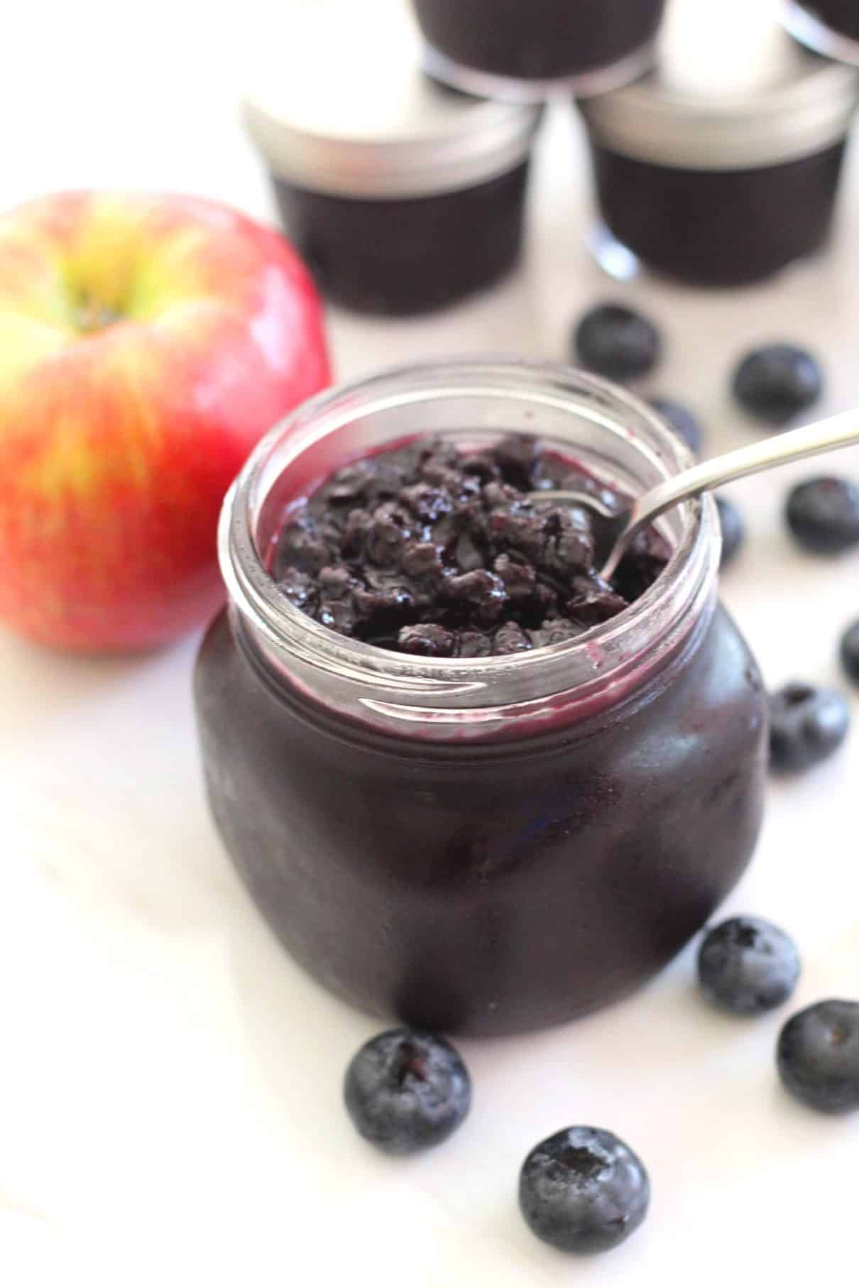 A spoon coming out of blueberry apple jam jar full of jam. In the background you see an apple and jars of jam!