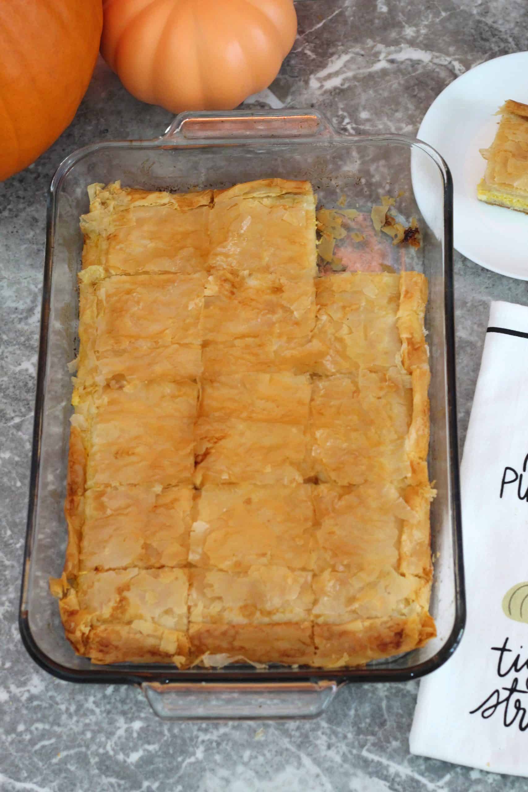 A baking tray with a phyllo baked dish, with a pumpkin towel next to the tray. A slice has already been cut out of this tray. 