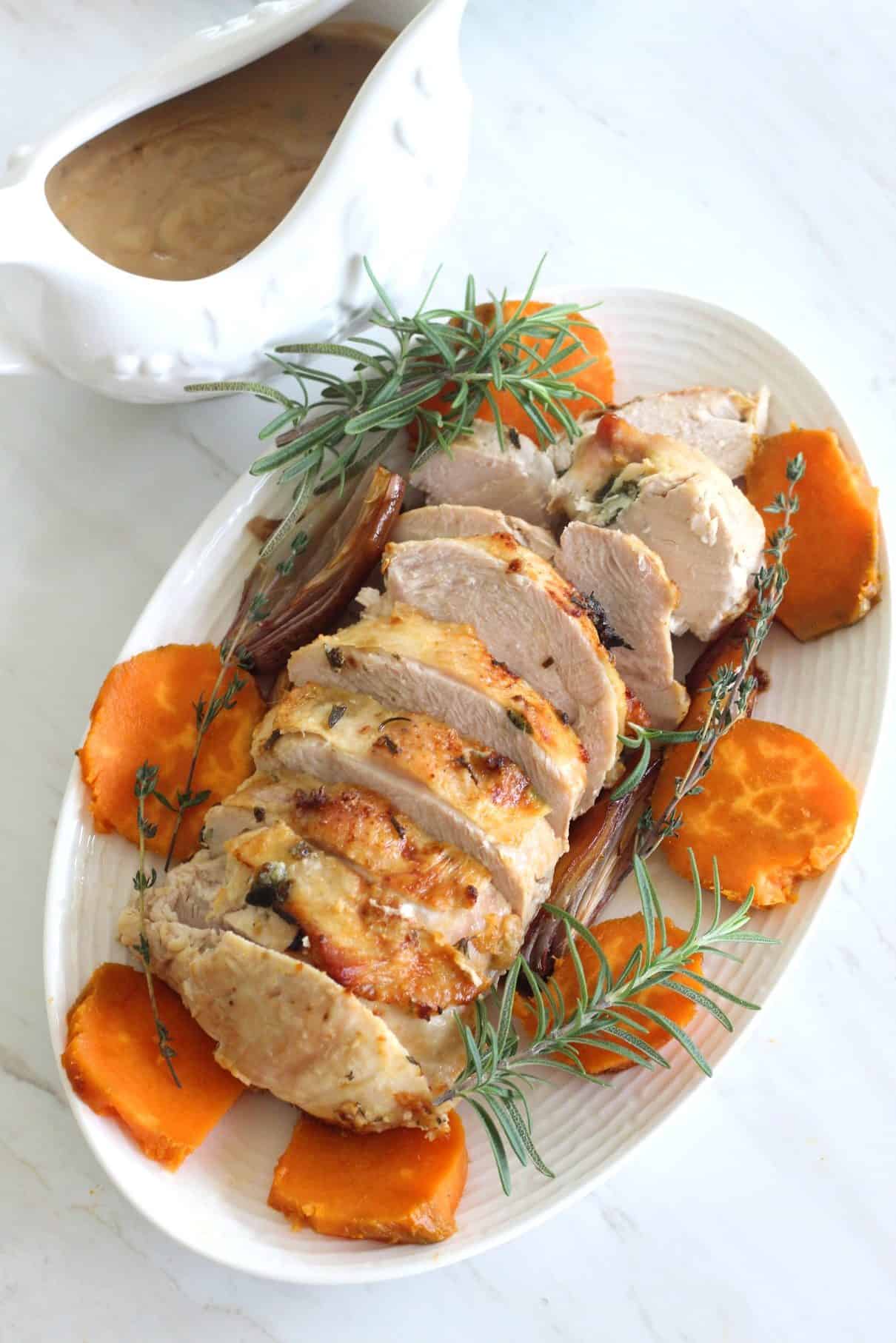 A platter with sliced roasted turkey, fresh herbs, sliced sweet potato and a side of gravy on the side. 