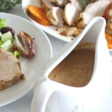 A gravy serving bowl with gravy in it, shown next to a plate of served food and another platter with sliced turkey.