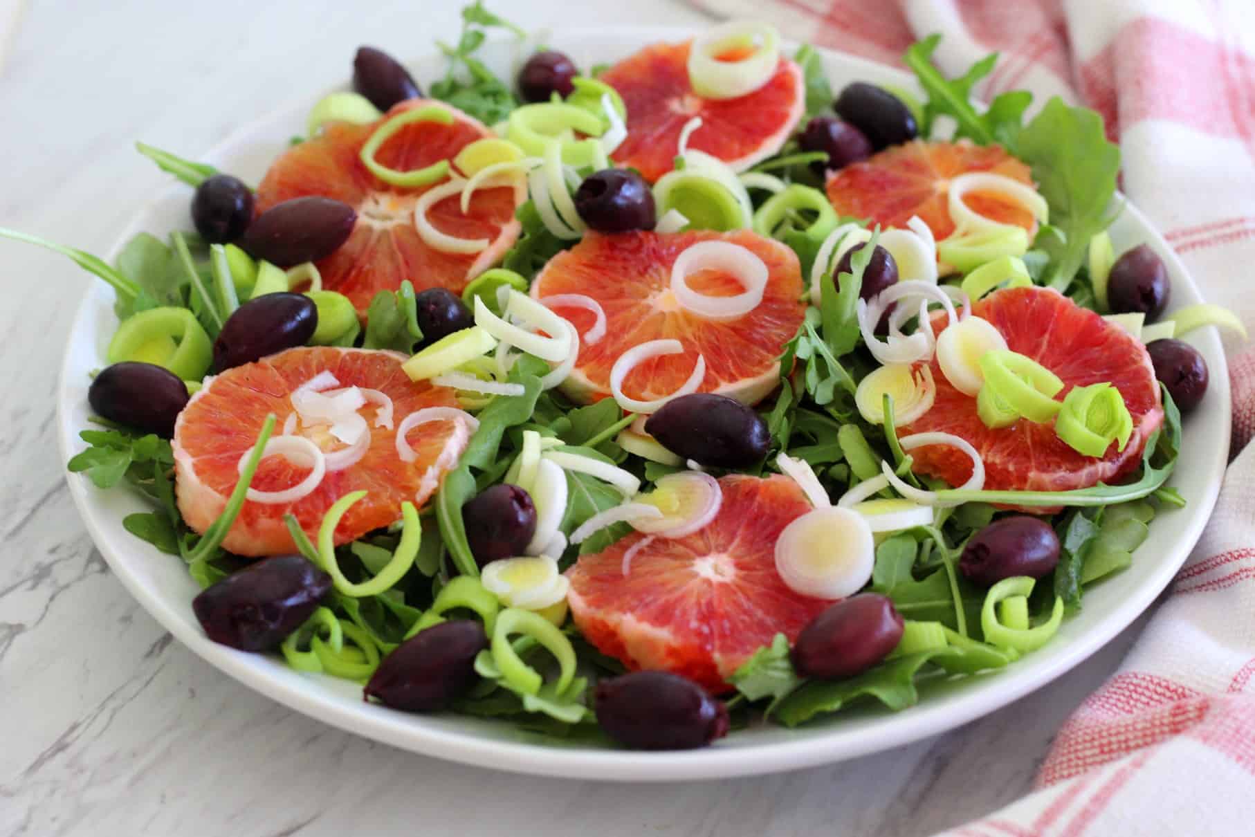 An orange salad, shown sideways. Ingredients visible on the salads are olives, leeks, greens and blood oranges sliced in circular shape.