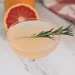 Fizzy pink looking cocktail in a coupe glass, garnished with rosemary. In the background there's half a grapefruit.