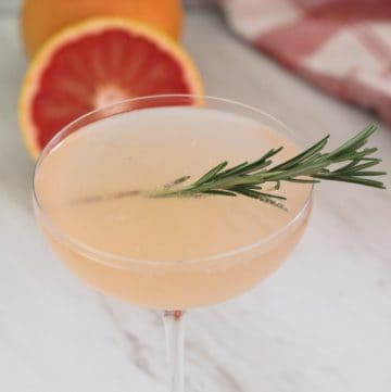 Fizzy pink looking cocktail in a coupe glass, garnished with rosemary. In the background there's half a grapefruit.