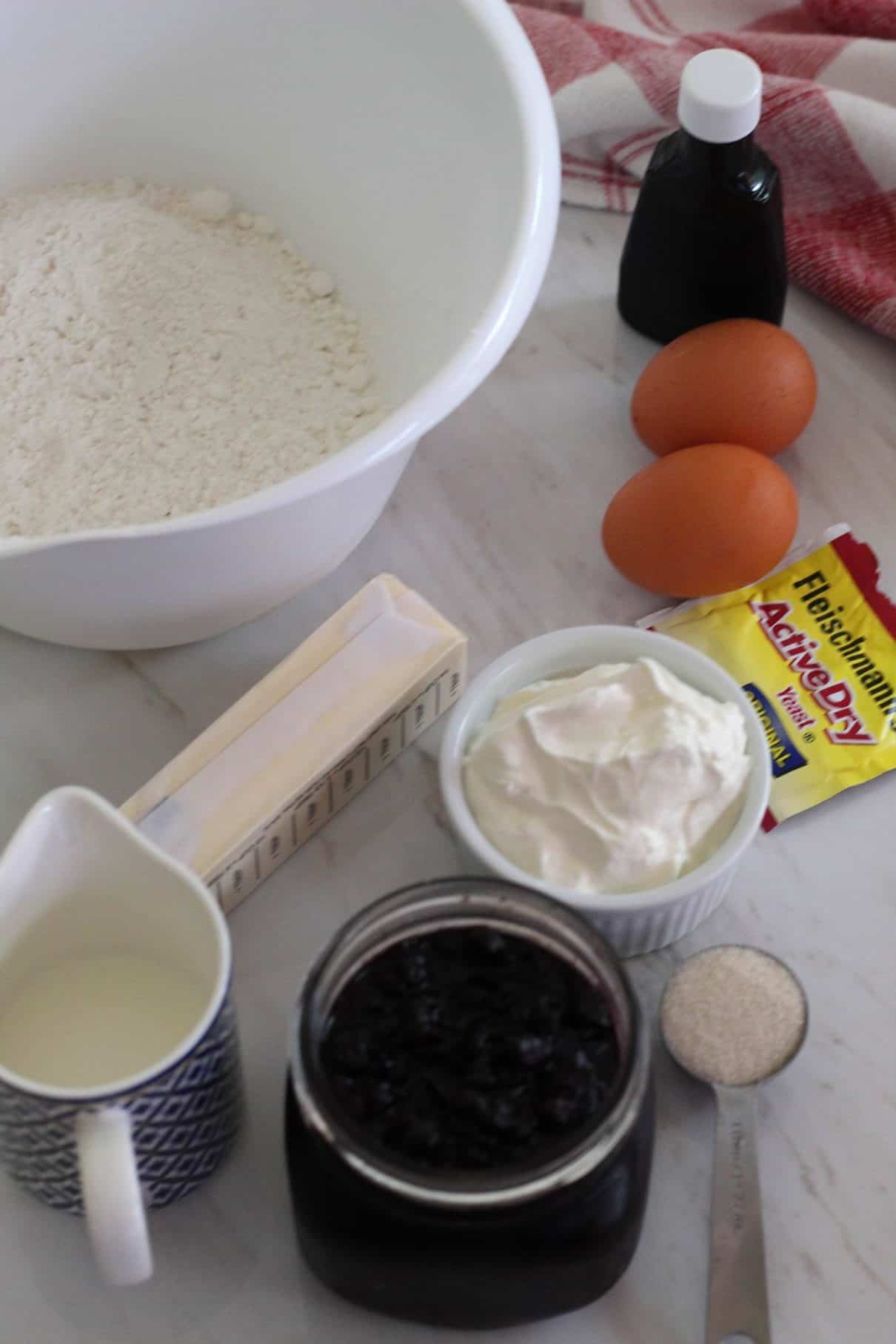 Several ingredients laid out for making the sour cream cookies. Flour, vanilla, eggs, yeast, sour cream, milk, sugar, butter and jam.