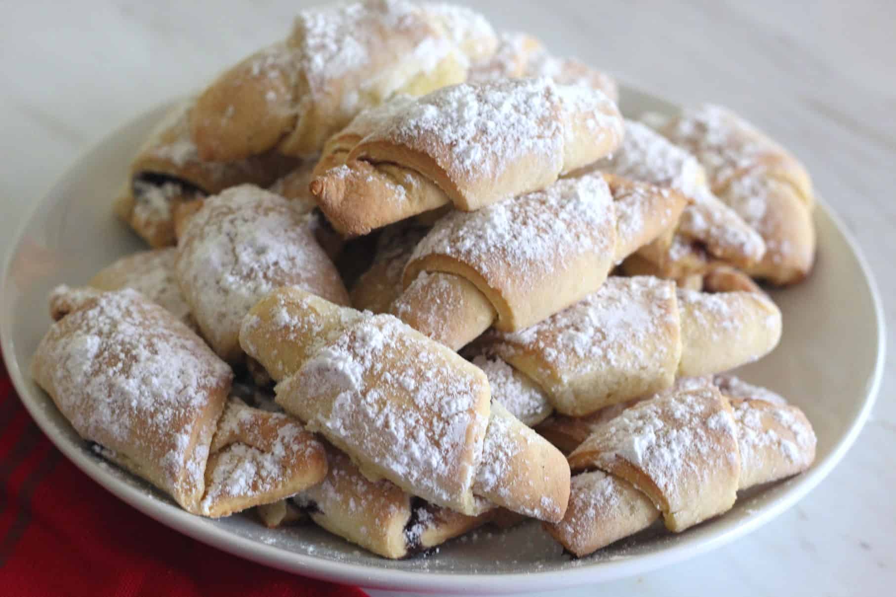 A plate with lots of rolled up crescent shape cookies that seem filled with jam.