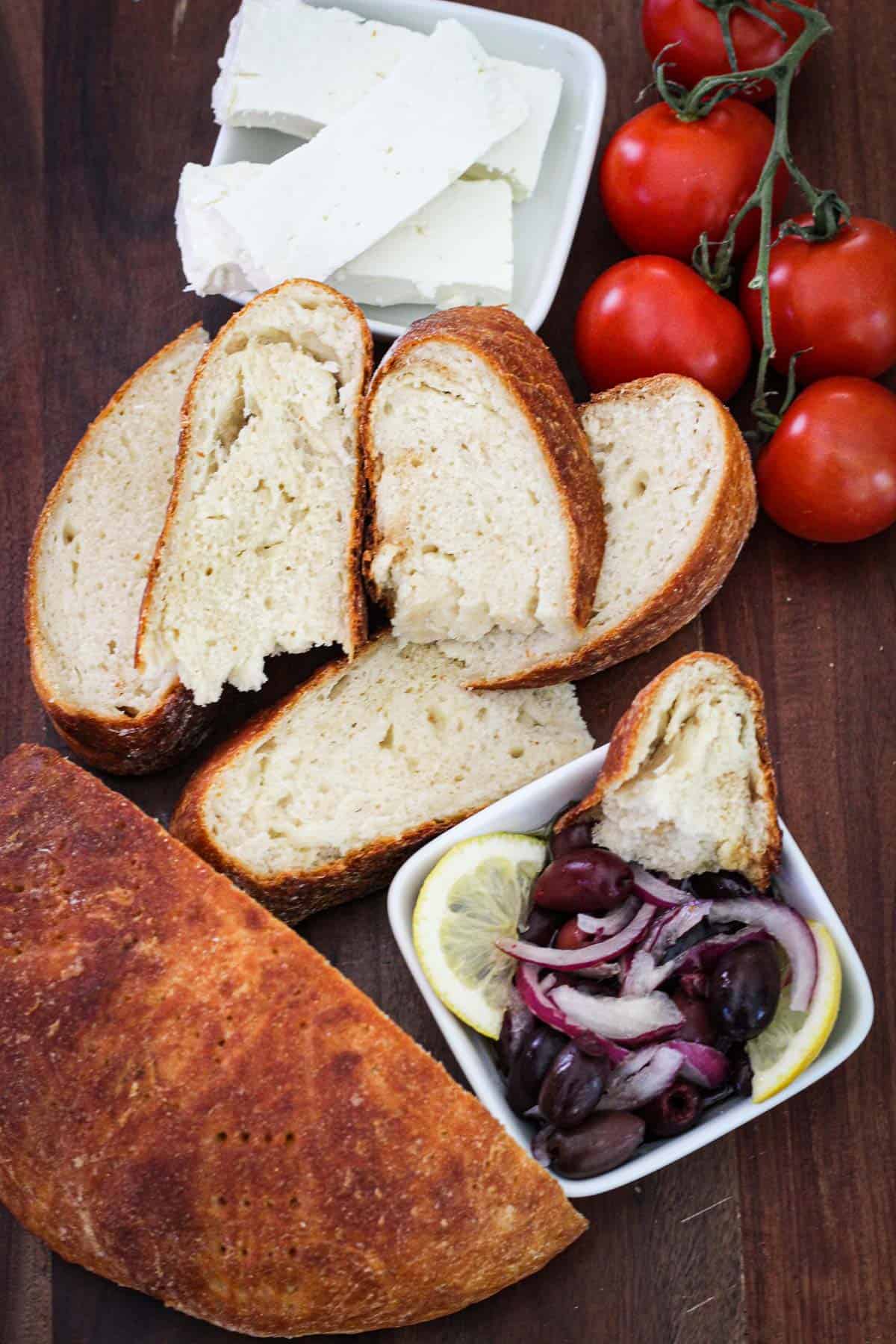 A cutting board with Albanian bread on it. Bread is cut in half, half is untouched in the corner and the other half is cut in slices. Then around the bread you see an olive appetizer, feta cheese and tomatoes.