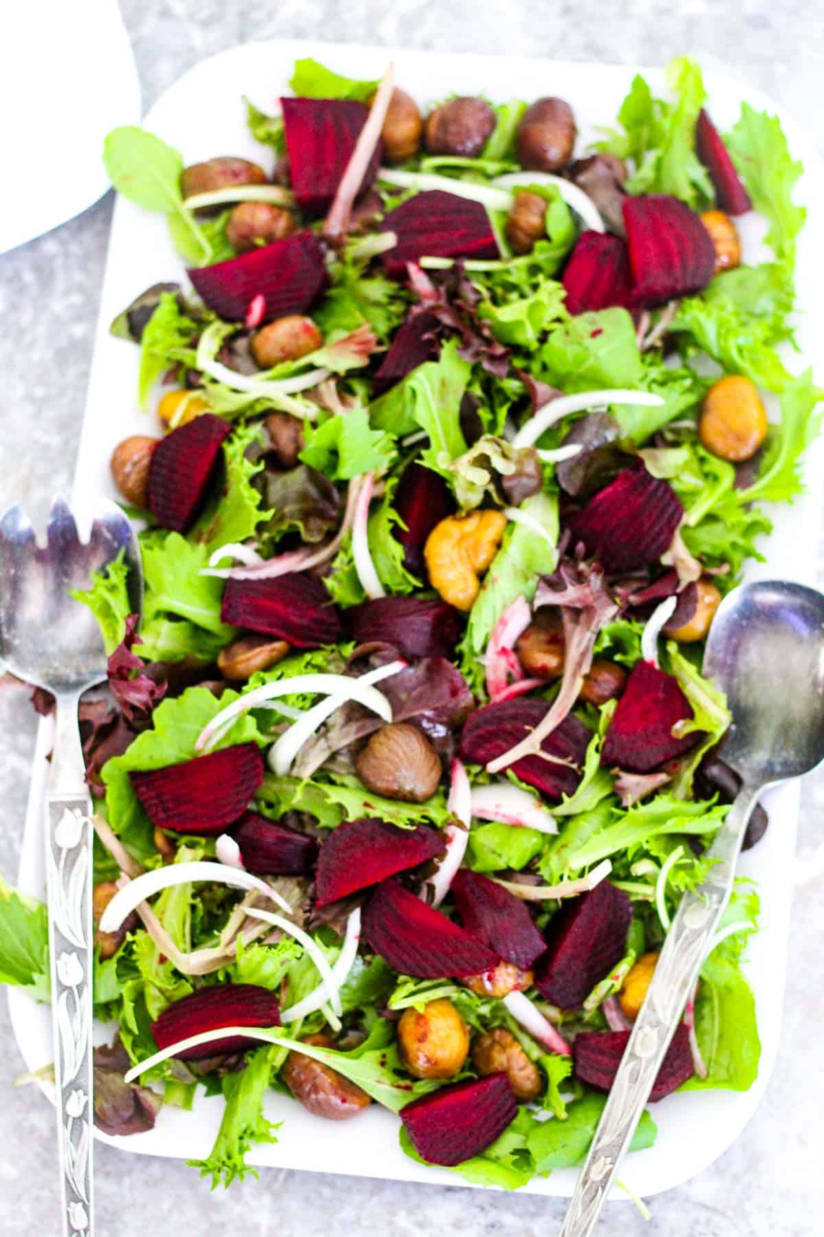 A rectangle shallow dish with a green salad that notably has beets, chestnuts and onions in it. Platter has 2 serving utensils on the side.