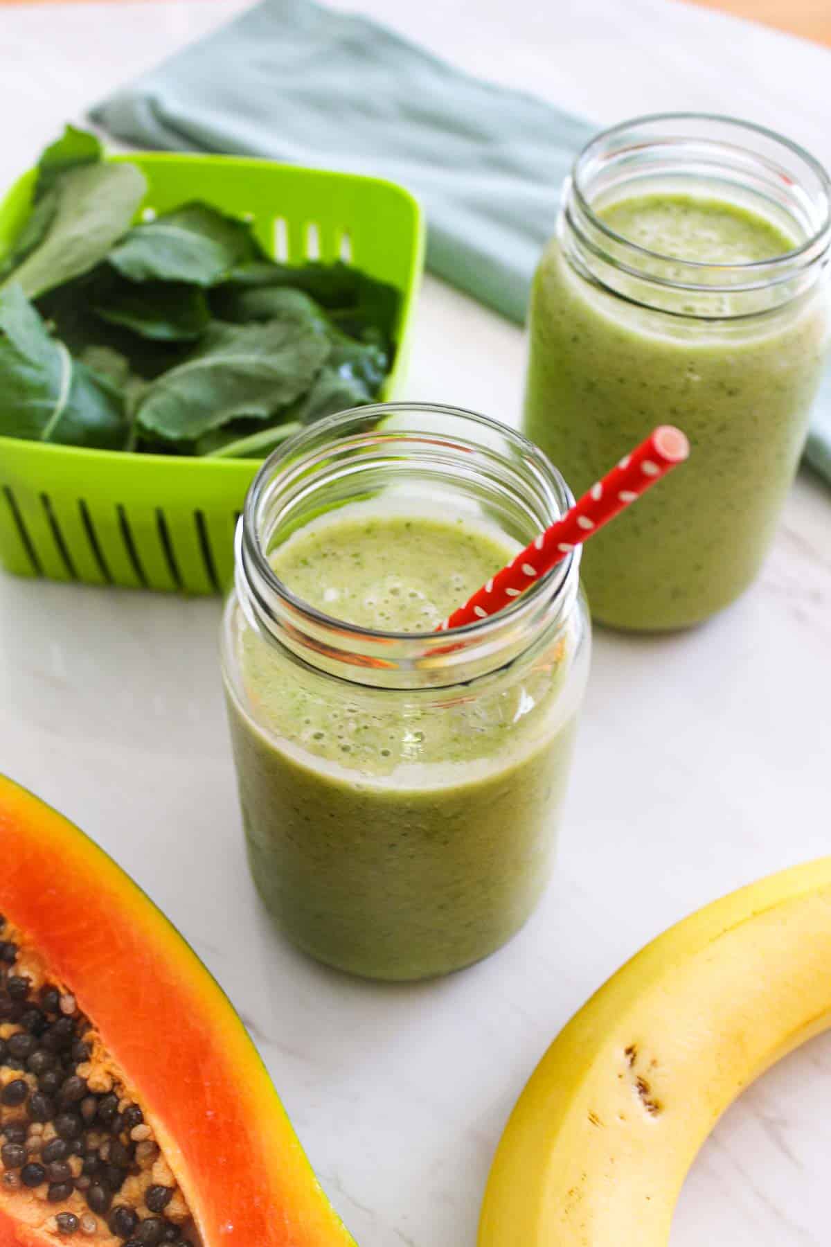 A jar of green smoothie, next to fresh kale, another jar of same smoothie, papaya and banana.