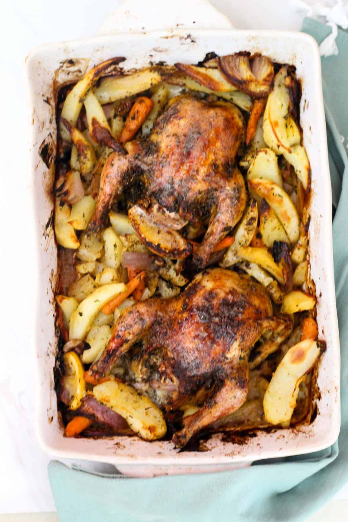 Roasted cornish hens and potatoes in a baking dish, there are 2 hens on the dish surrounded by lots of potatoes.