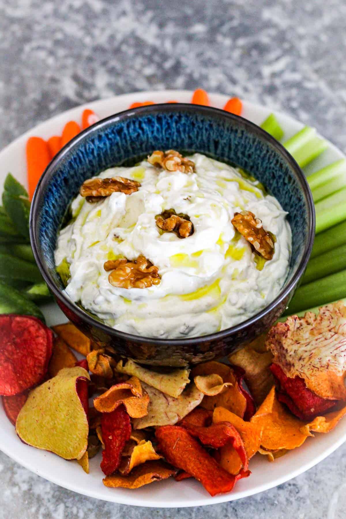 A bowl of sour cream dip with walnuts and garlic, garnished with walnuts and olive oil. Dip is served with colorful chips and vegetables.