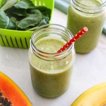 Picture focuses on a jar of green smoothie, next to it there's another jar of same smoothie without the straw, fresh kale, papaya and a banana.