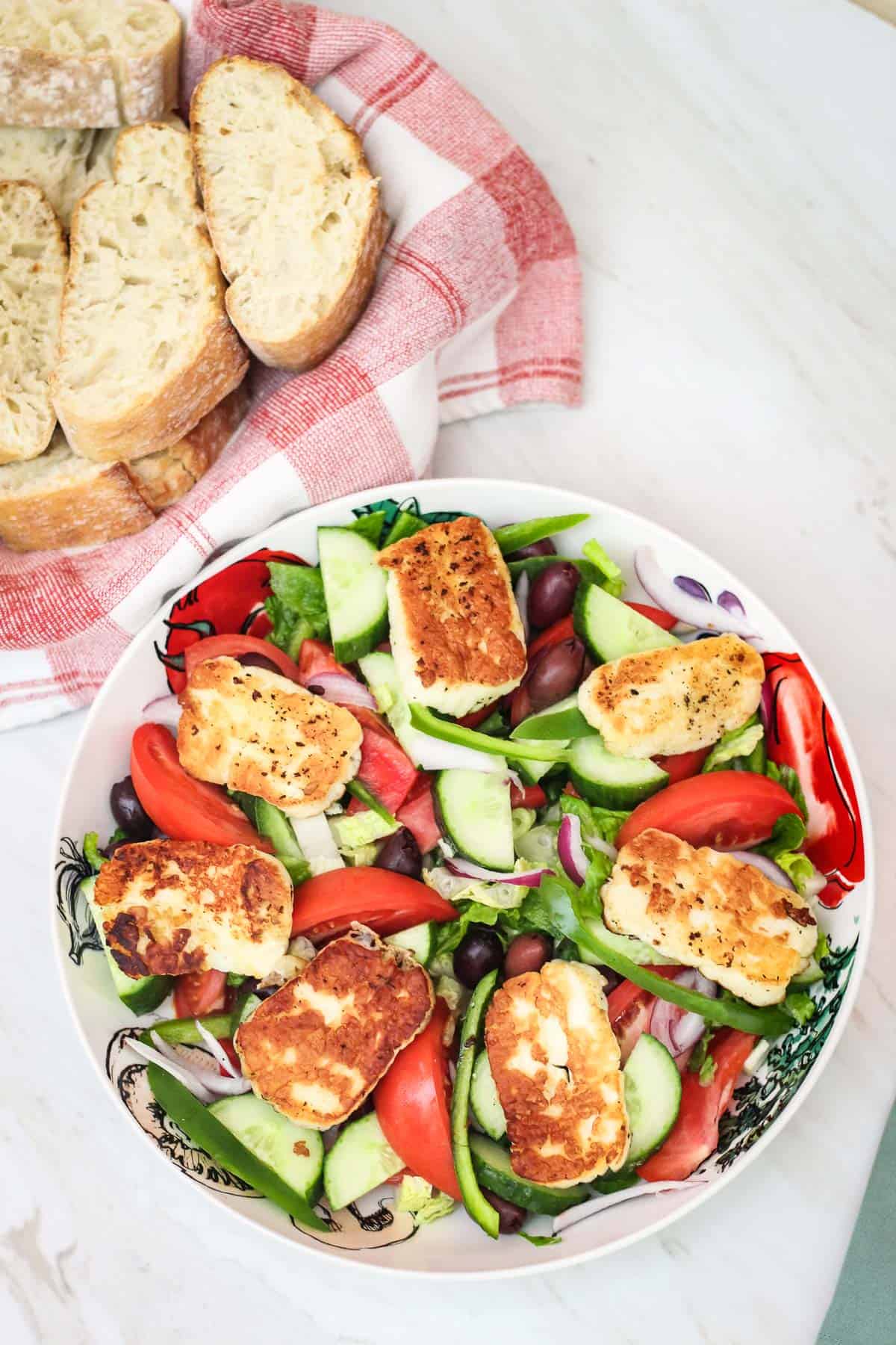 A platter of big family style salad topped with fried halloumi cheese shown next to a basket of fresh bread.
