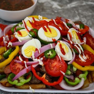 A platter with fries, meat, vegetables, eggs and sauces shown next to a large bowl of salsa.