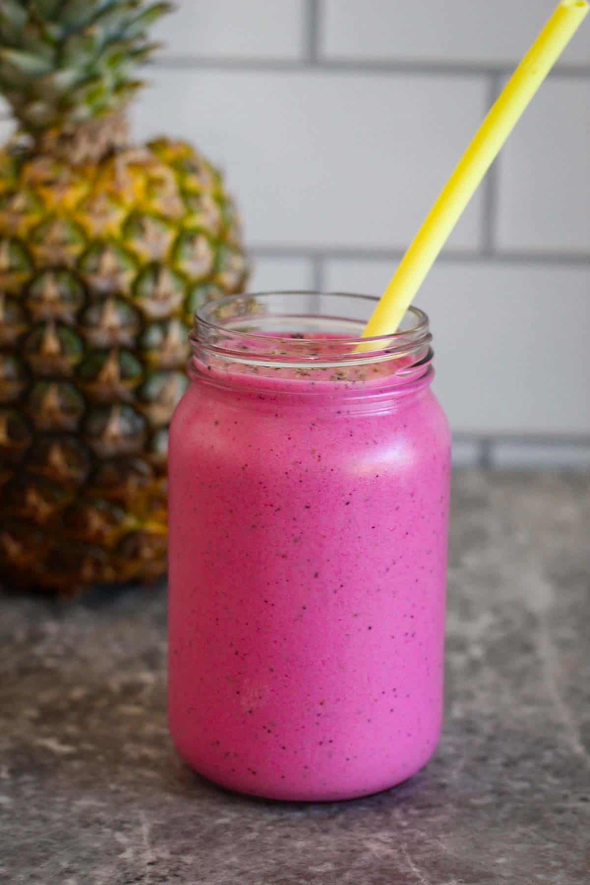 A jar of colorful smoothie, with a straw in it. There's a pineapple in the background while the jar is front and center.