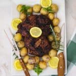A roasted lamb leg on a platter garnished with potatoes, rosemary, mint, lemon etc.