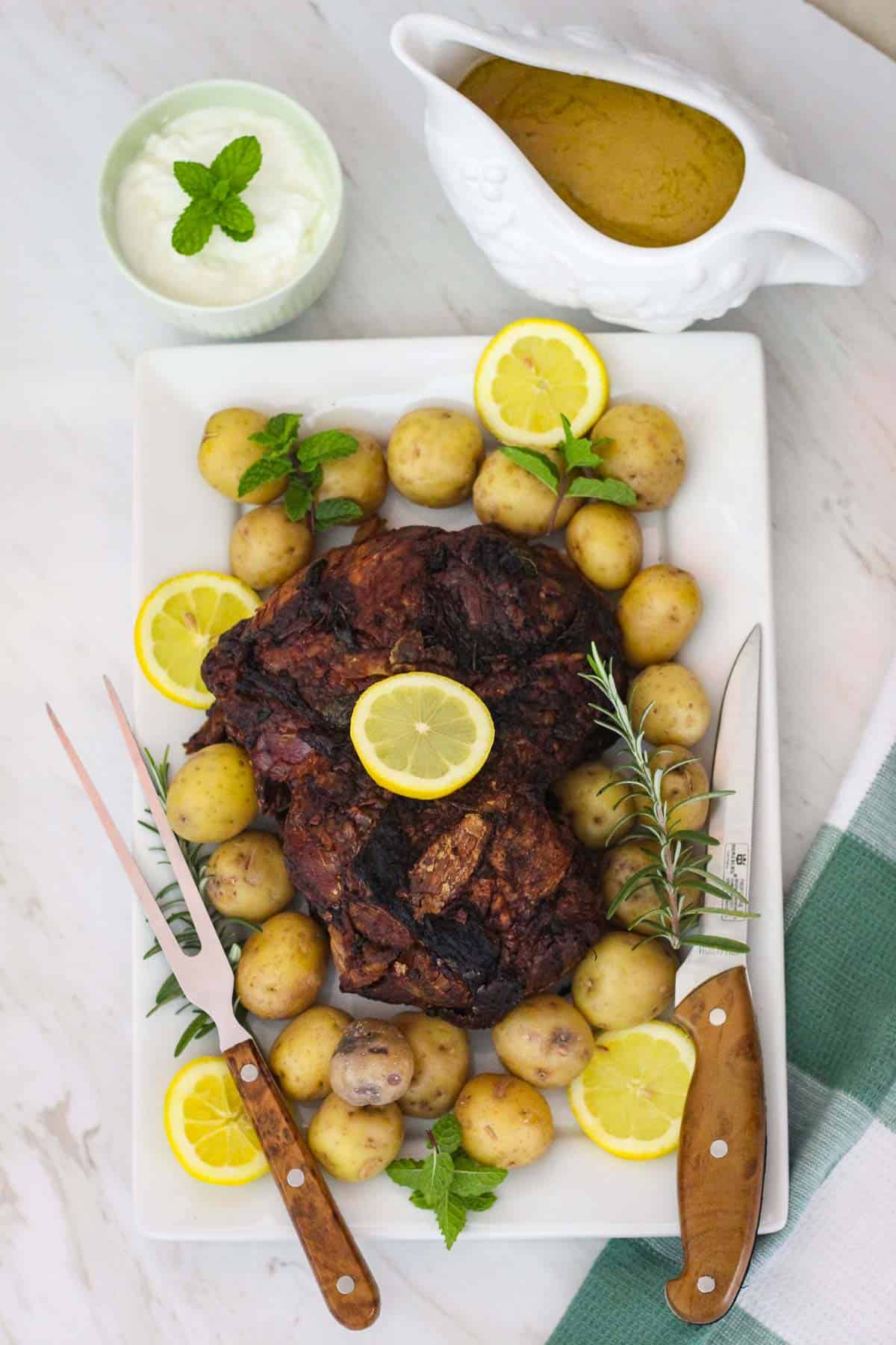 A platter with roasted lamb leg, potatoes, rosemary, lemon, cutting utensils, mint. Next to the platter there's a gravy jar and a yogurt dip.