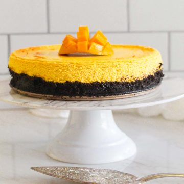 A mango cheesecake with oreo crust on a cake display. There's fresh mango cut on top of the cheesecake and a serving utensil