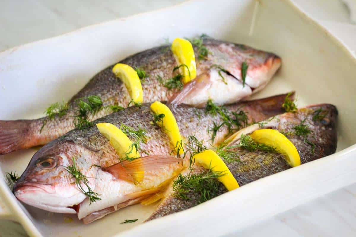 Red snappers ready to bake in the baking dish. There are slits cut through the snapper where lemon slices are fit in. There's also lots of fresh dill over the fish. 