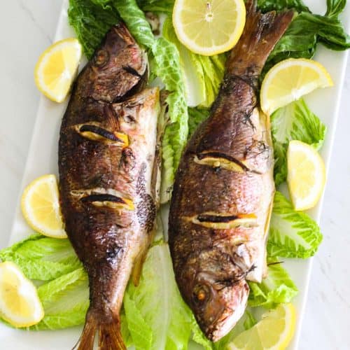 2 whole baked fish, Red Snapper on a platter with greens and lemons.