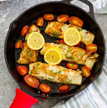 Cast Iron cooked halibut shown right after cooking. Skillet is shown with 4 halibut fillets, garnished with lemon slices, diill and cherry tomatoes.