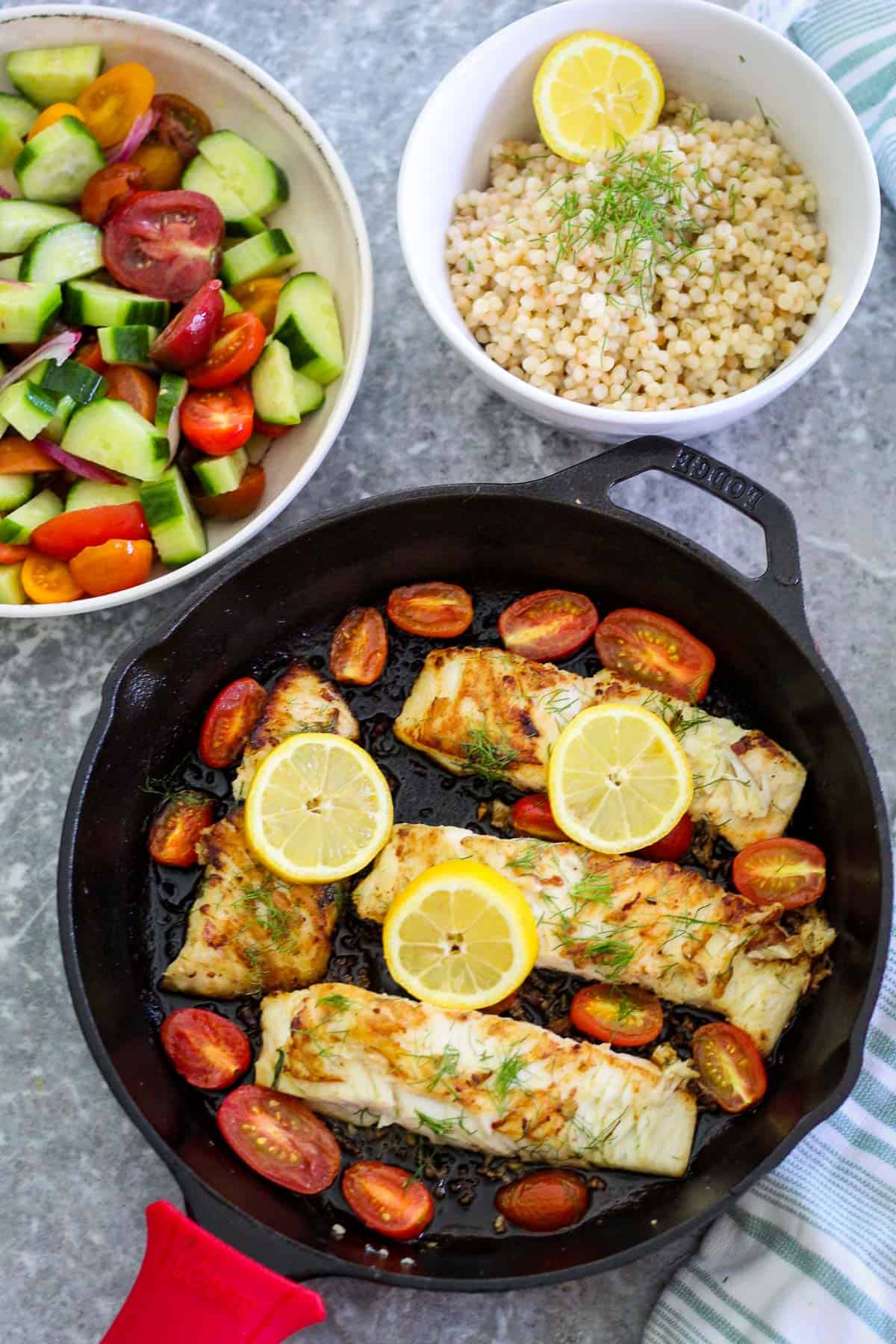 Halibut fillets in a cast iron skillet, shown with cherry tomatoes and lemon. There's a salad and couscous dish on the side as side dishes. 