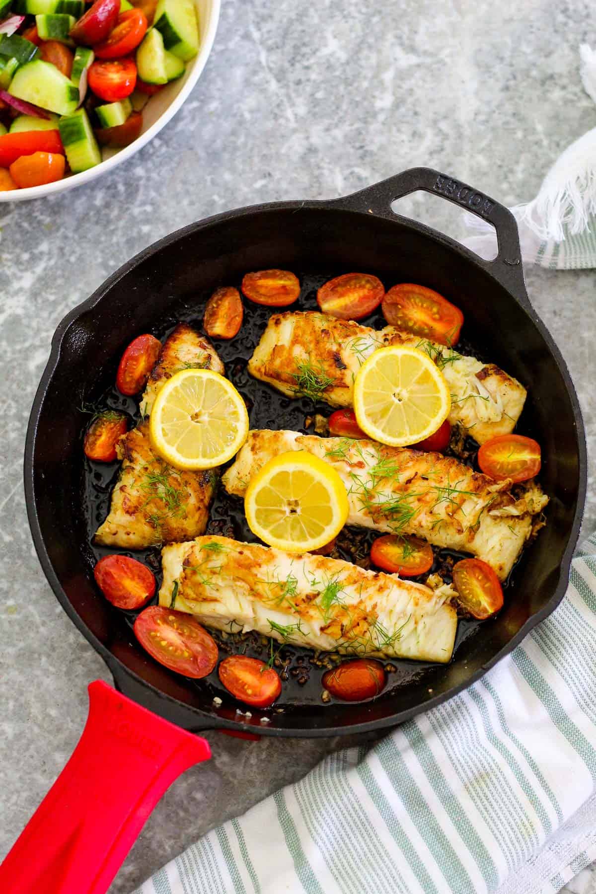 Seafood dinner with halibut fish - this dish is shown cooked on a cast iron skillet. There are 4 fish fillets in the skillet, lemon slices, cherry tomatoes and everything is garnished with dill. 