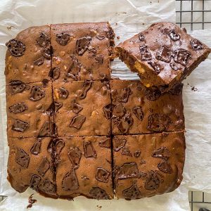 Picture shows just baked and cut brownies on a cooling rack. Brownies have been cut into a 9 pieces in a 3x3 format, one piece is slightly slanted so you can see the slice sideways.