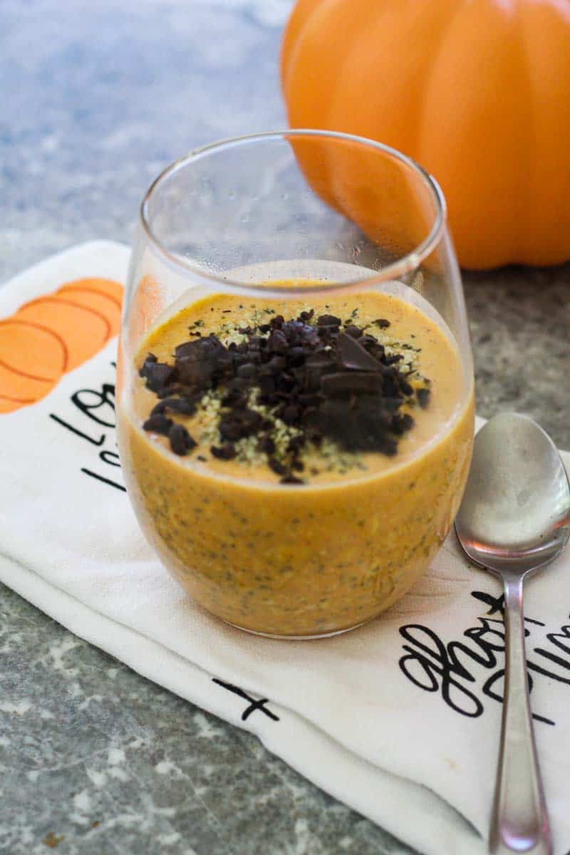 A clear glass, filled halfway with pumpkin chia oatmeal and topped with shaved dark chocolate. Glass is over a pumpkin themed kitchen towel and there's a spoon next to it.