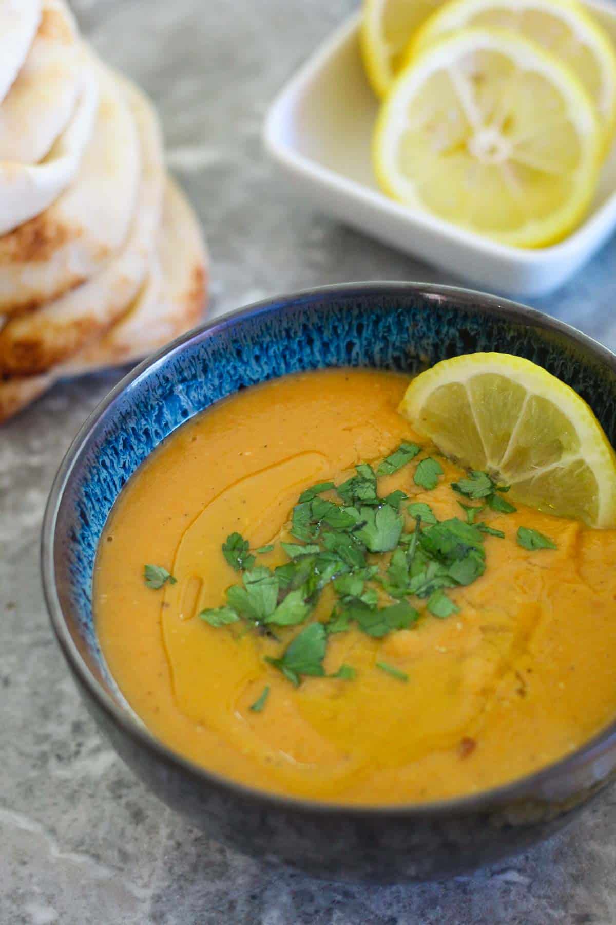 A bowl of Red Lentils Soup with Butternut Squash, garnished with parsley and lemon. In the background you see a stack of pita breads and lemon slices.