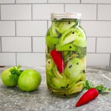 There's a big closed jar with pickled green tomatoes, sliced. You can see a red chili and some peppercorn in the jar too. Next to the jar there's 2 whole green tomatoes, parsley and a red chili.