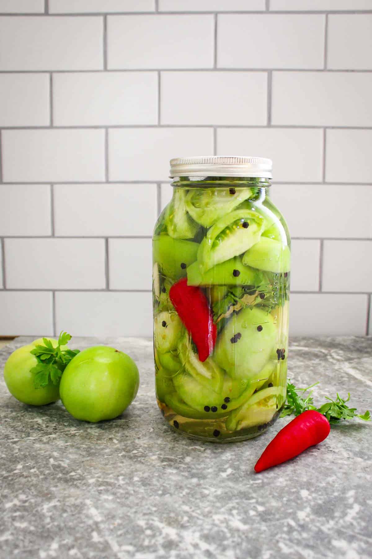 A jar of sliced, pickled green tomatoes. There's peppercorn throughout the jar and a red chili pepper. 