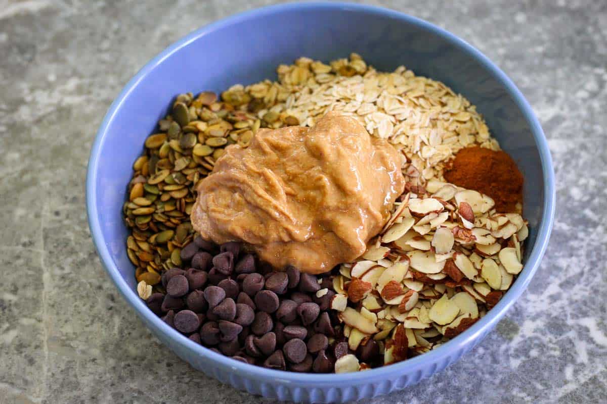 A blue bowl full of ingredients, not mixed yet. You can see on a circular shape oats, almonds, chocolate chips, cinnamon, pumpkin seeds and peanut butter.