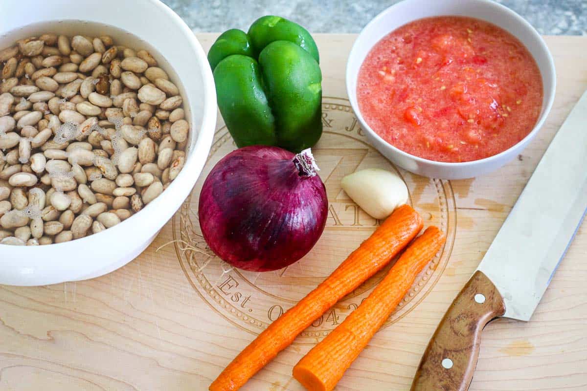 Fresh ingredients for the soup: pinto beans in the water, green pepper, blended tomatoes, red onion, garlic, carrot on a cutting board with knife. 