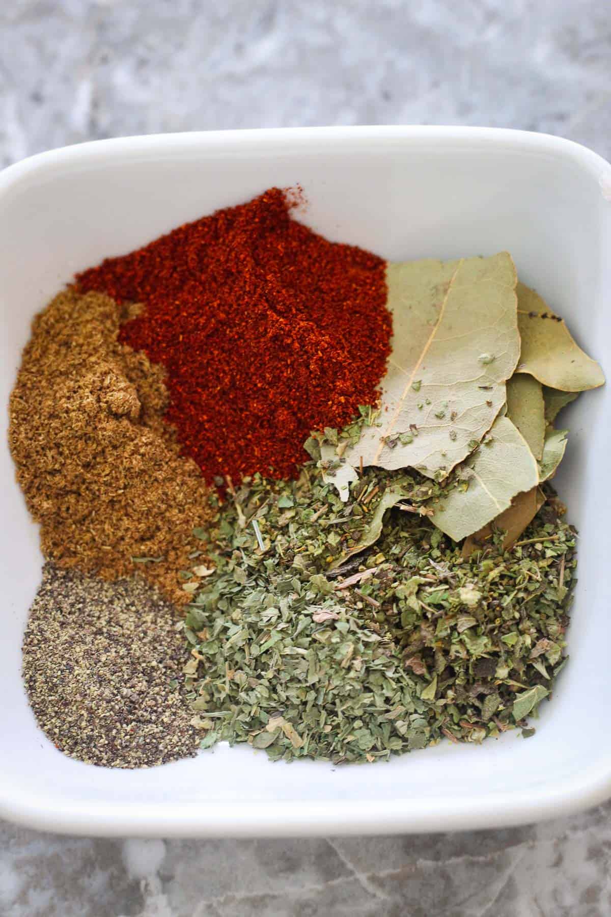 Seasonings for pinto beans soup in one square bowl: bay leaves, smoked paprika, cumin, black pepper, parsley and oregano.
