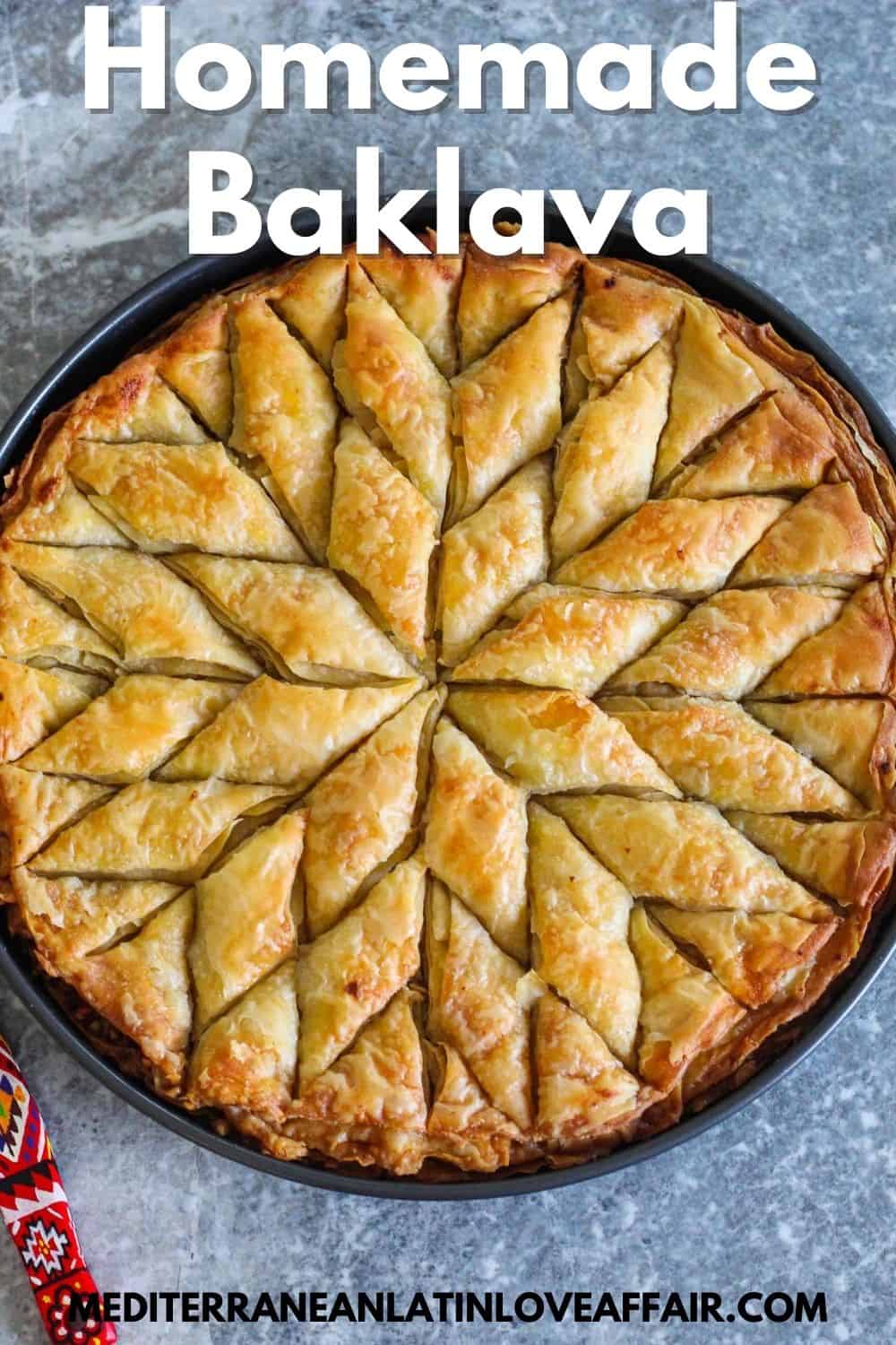 An image prepared specifically for Pinterest. It shows the baked baklava in a baking tray, round shaped. Baklava is cut in diamond shape. There's a title bar over the image that says Homemade Baklava and there's a website link at the bottom of the image. 