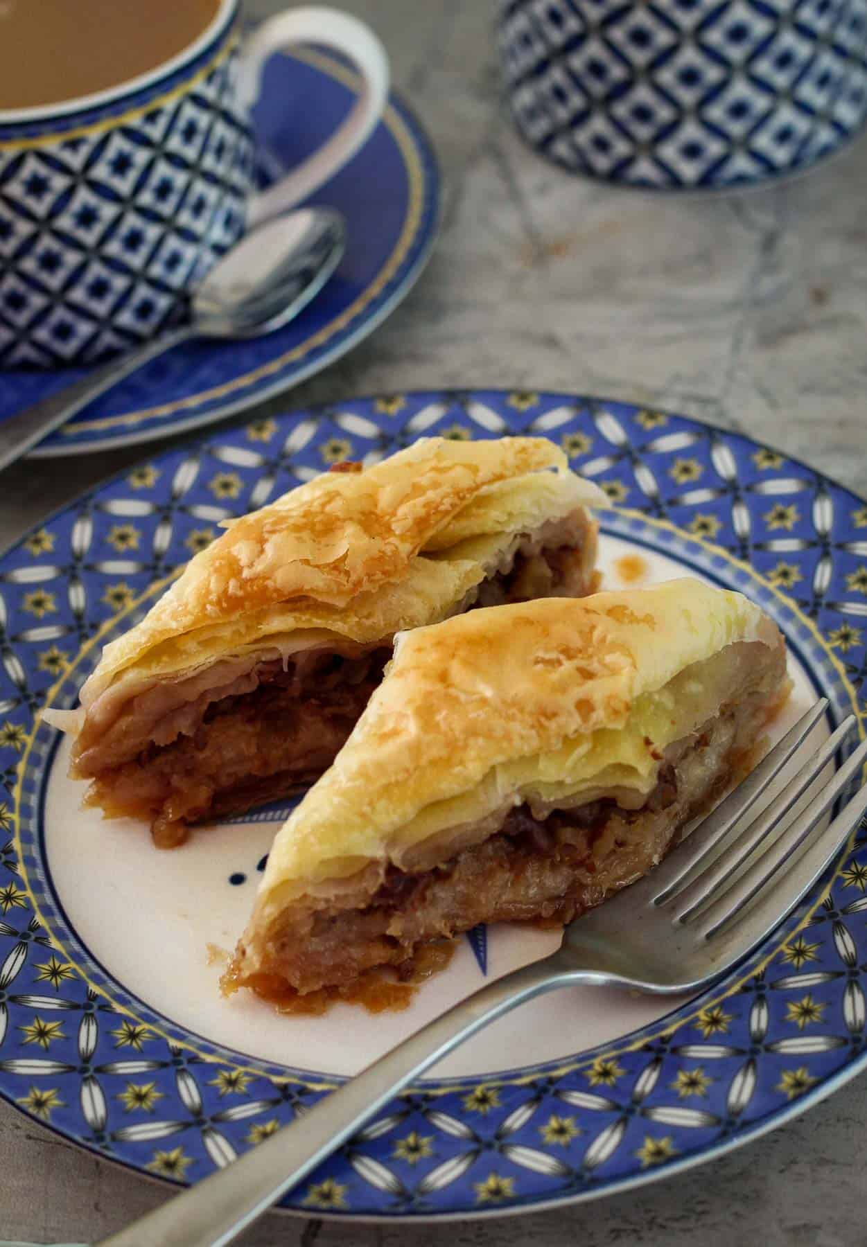 A plate with 2 slices of baklava and a fork. On the background there's a partial view of a cup of coffee.