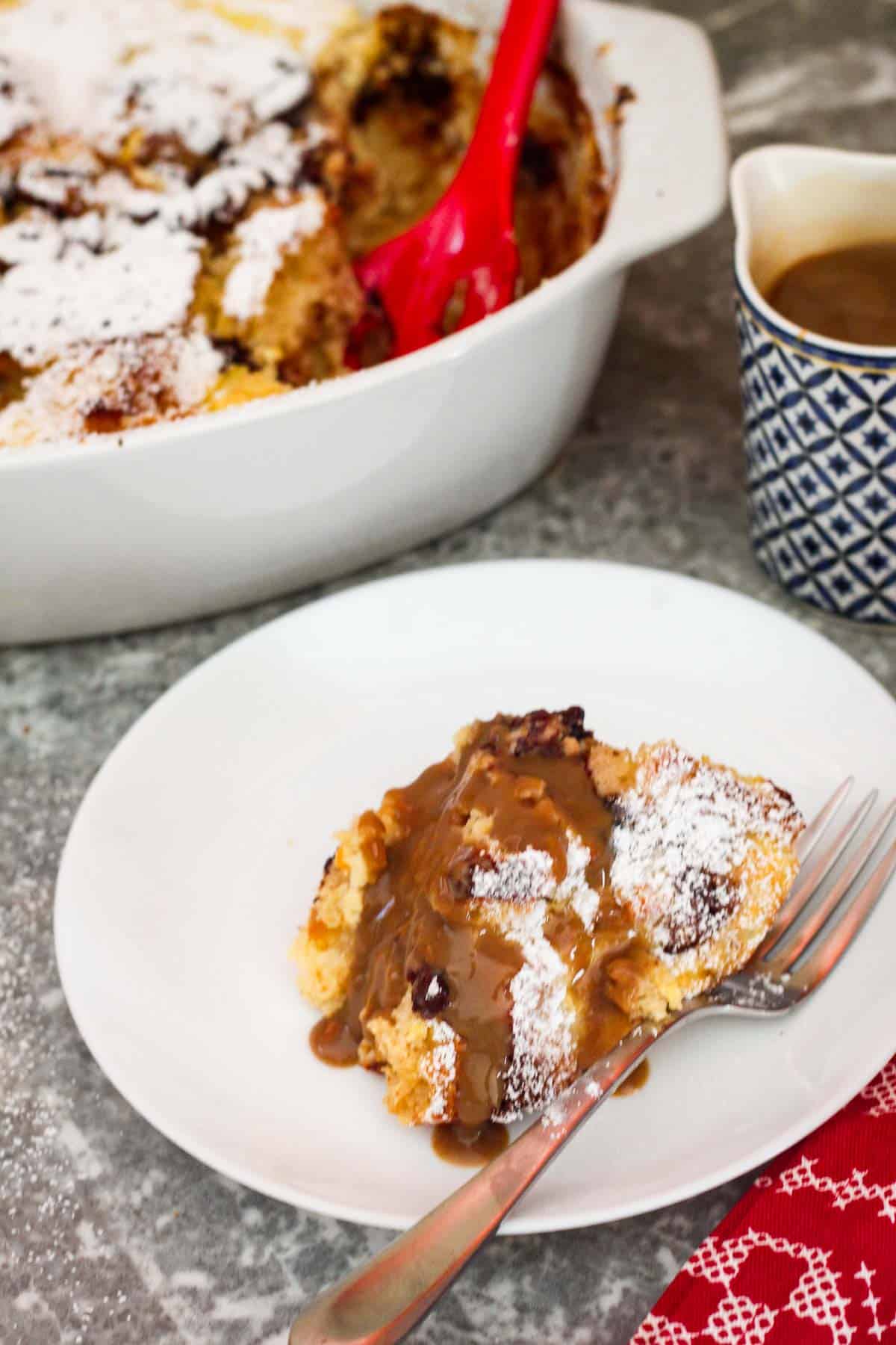 Serving of panettone bread pudding drizzled with kahlua cream sauce. The whole baking dish with the pudding is in the background as well as the serving container for the kahlua sauce. 