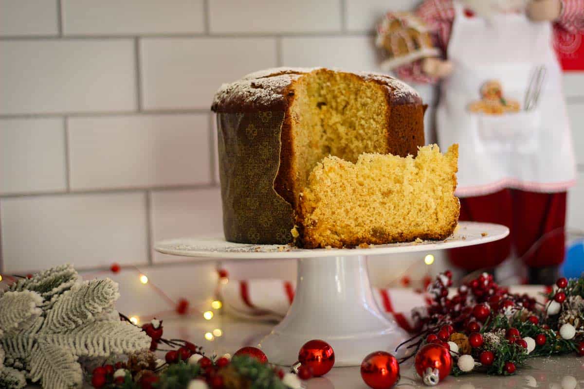 A sliced panettone on cake holder, shown with a slice down and the rest of the panettone up. There's a festive atmosphere around the panettone with lots of decorations and lights.