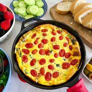 A frittata brunch: cast iron skillet with frittata in the middle, than surrounding the skillet you see bread, cucumbers, olives, fruits, salad etc.