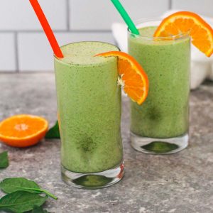 Green smoothies made with matcha, spinach and mandarins. There are 2 smoothie glasses, garnished with fresh mandarin slices. There's spinach on the counter and each smoothie glass has a different colored straw.