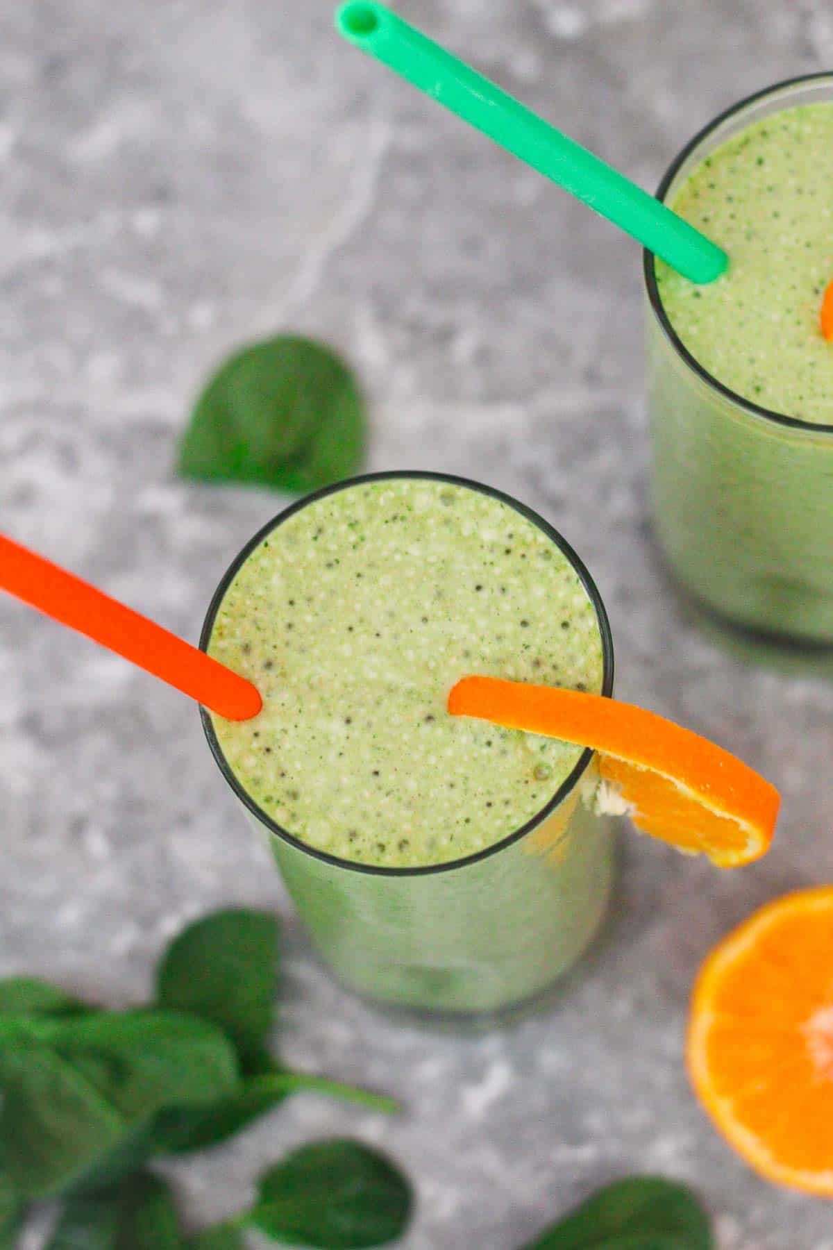 Matcha, Spinach and Mandarin smoothies. Picture shows 2 smoothie glasses from overhead angle, one glass in the center, one in the background. Glasses are garnished with fresh mandarin slices. 