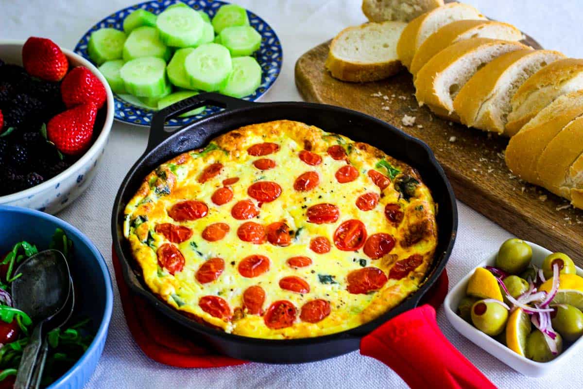 A brunch setup with Mediterranean frittata in a cast iron pan surrounded by side dishes and bread.