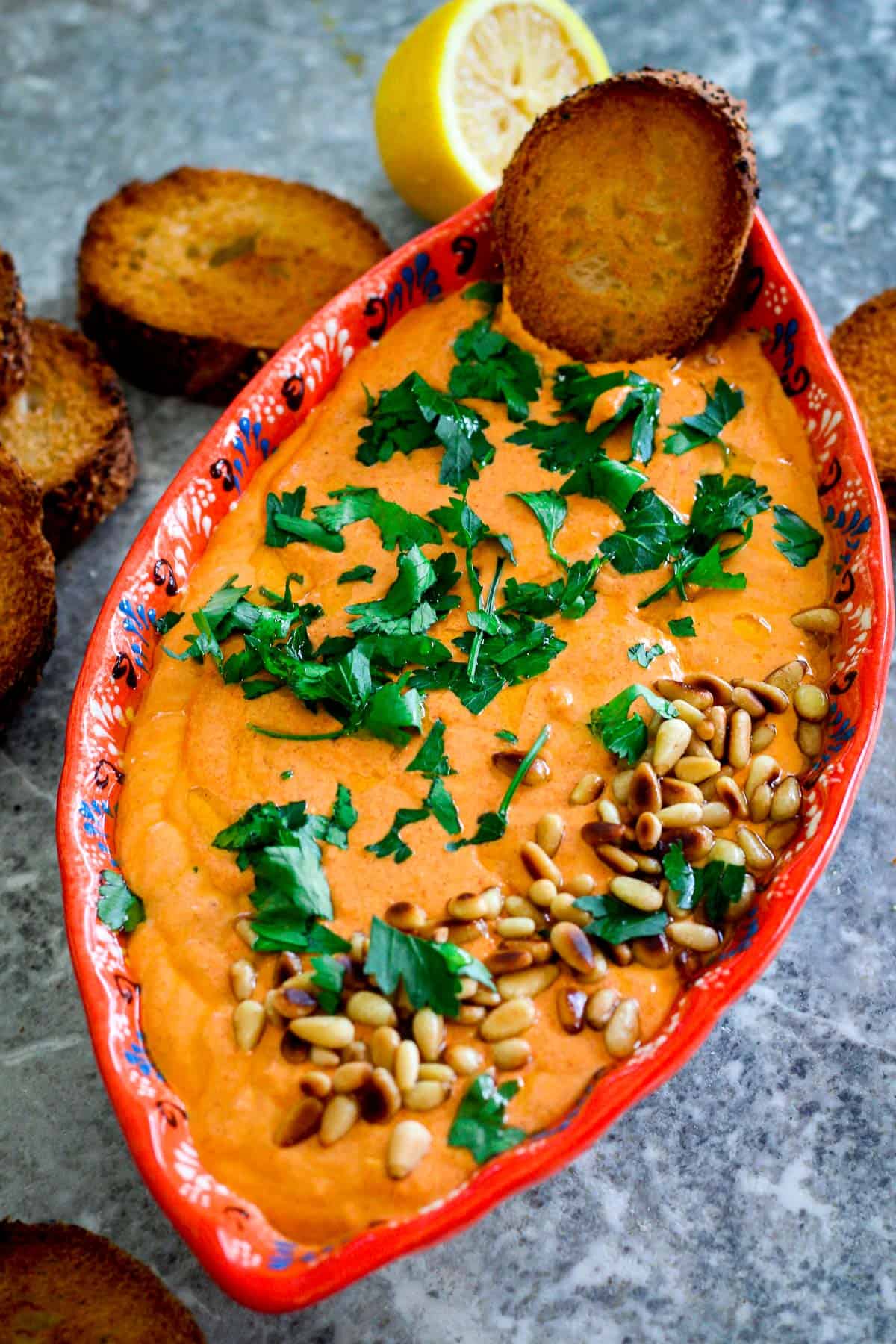 A colorful platter with a creamy orange looking dip made with red peppers and feta cheese. Dip is garnished with pine nuts and fresh parsley and there's a bread dipped in it!! 
