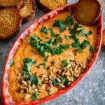 A colorful platter with a dip made with red peppers and feta. Dip is garnished with pine nuts and fresh parsley. There's toasted bread dipped in the platter and more bread on the side!!