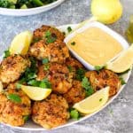 Baked fish cakes served on a platter with a dip, garnished with lemon slices and parsley.