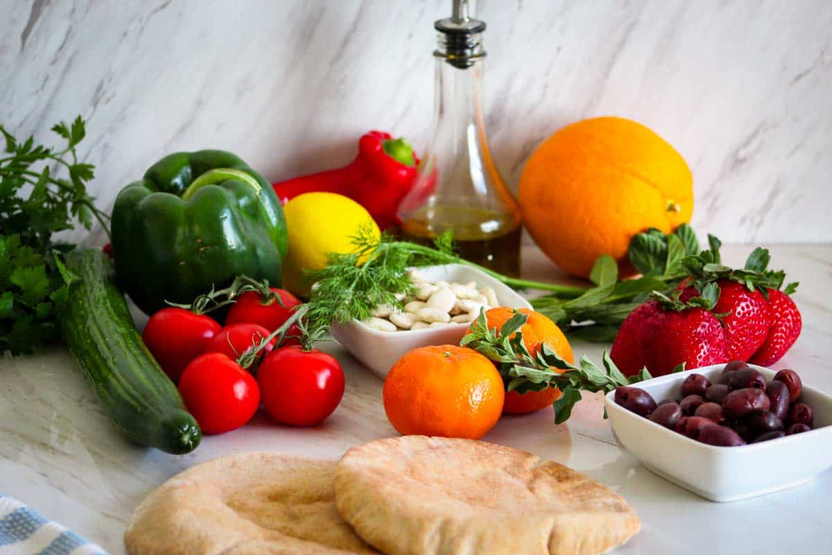 A set of grocery items on a counter, they look very vibrant and fresh. Some of the most notable items are tomatoes, cucumber, peppers, lemon, orange, strawberries, olives, mandarins, olive oil, beans and pita bread.