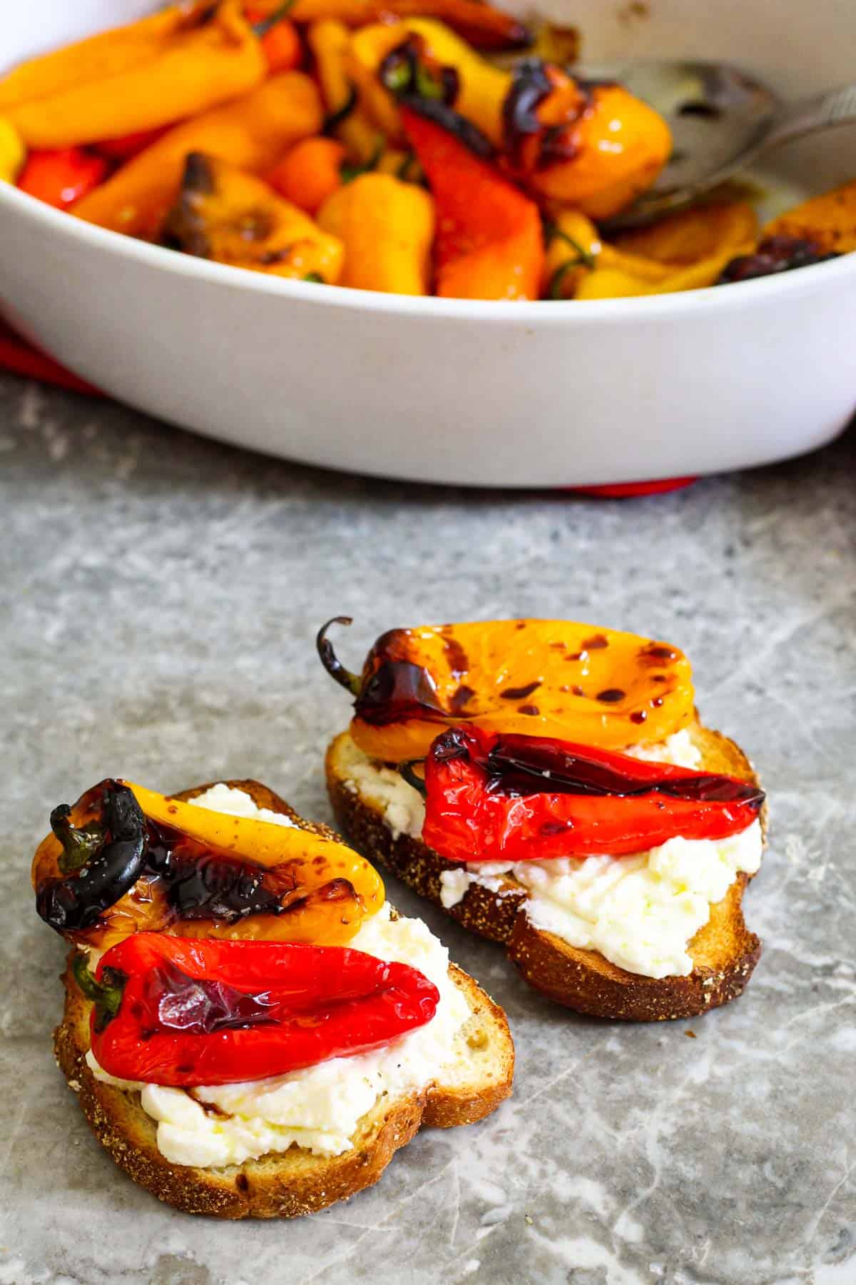 Toasted sourdough bread (2), topped with ricotta layer and over ricotta there's 2 roasted pepper on each toast. In the background, you see the baking dish for the peppers. 