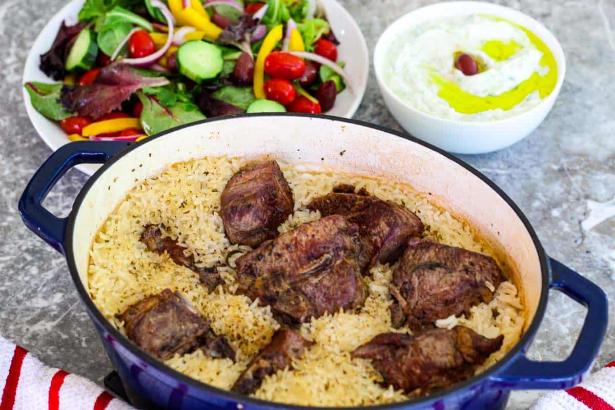 A blue dutch oven with a lamb/rice casserole shown next to a big, colorful salad and a tzatziki bowl.