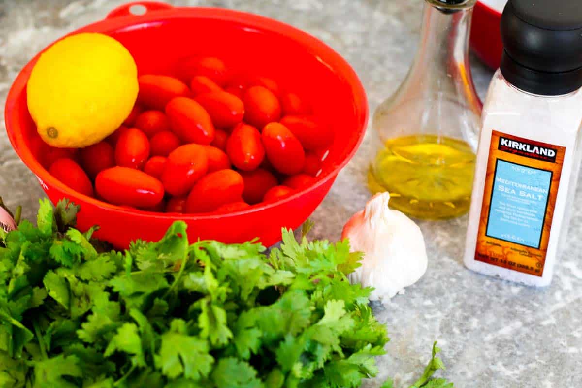Ingredients all together before marinating the tomatoes: cherry tomatoes on a colander with 1 fresh lemon, olive oil, Mediterranean sea salt, garlic head and a bunch of cilantro. 